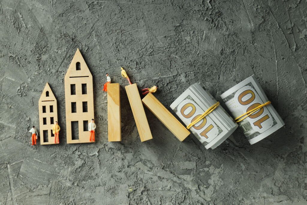 Small wooden house models toppling beside bundles of currency on a gray surface