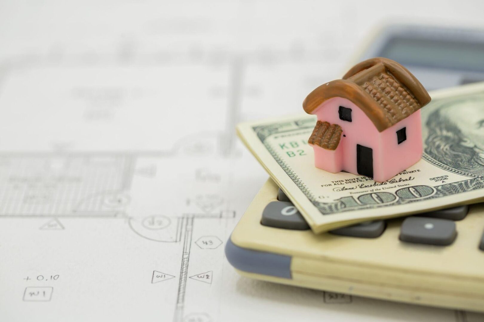 Miniature house on dollar bills placed over construction blueprints and a calculator