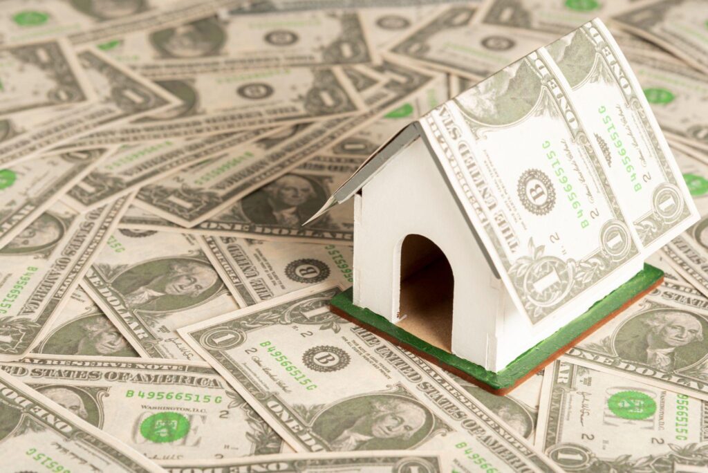 Small white model house covered in dollar bills placed on a surface full of U.S. currency