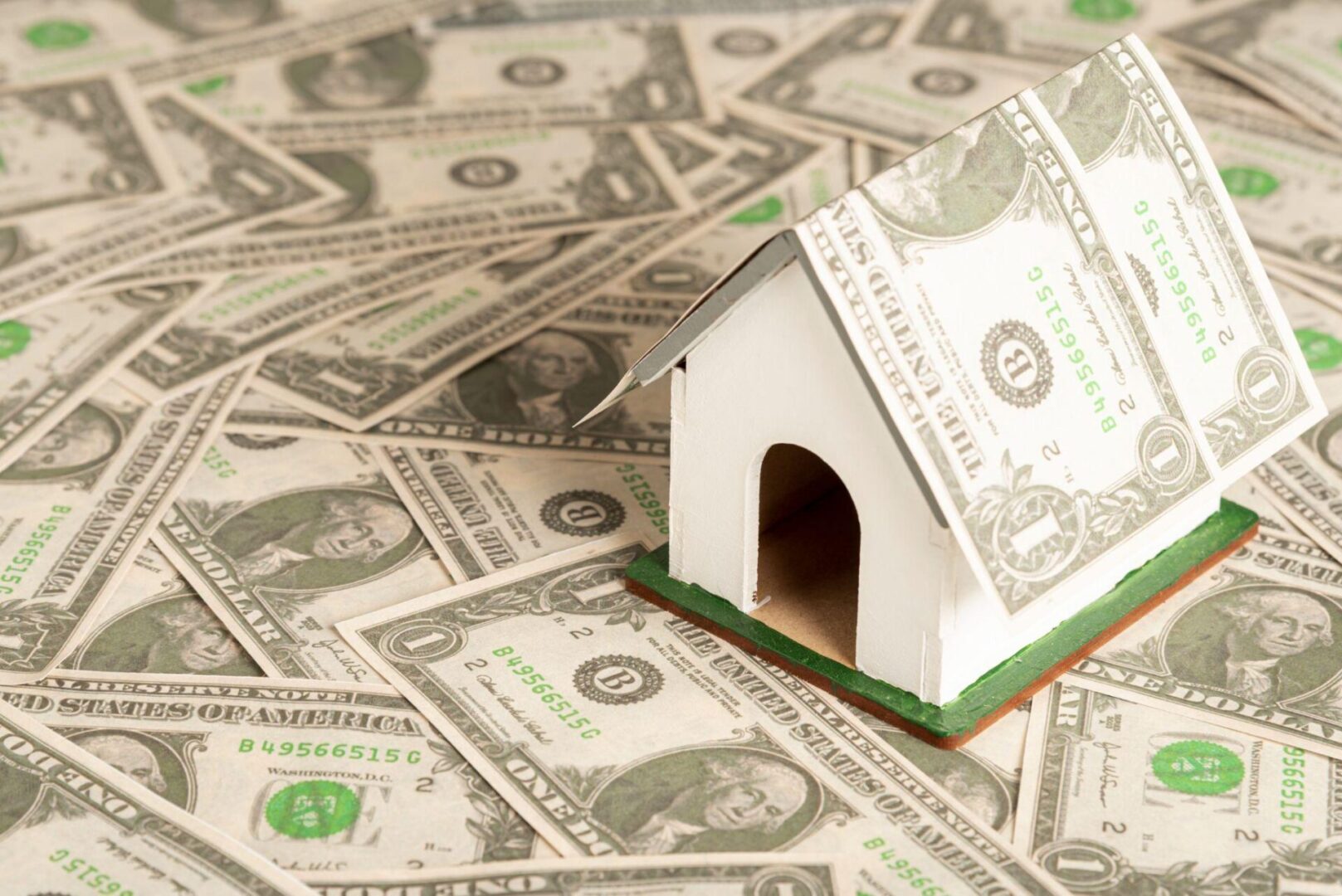 Small white model house covered in dollar bills placed on a surface full of U.S. currency