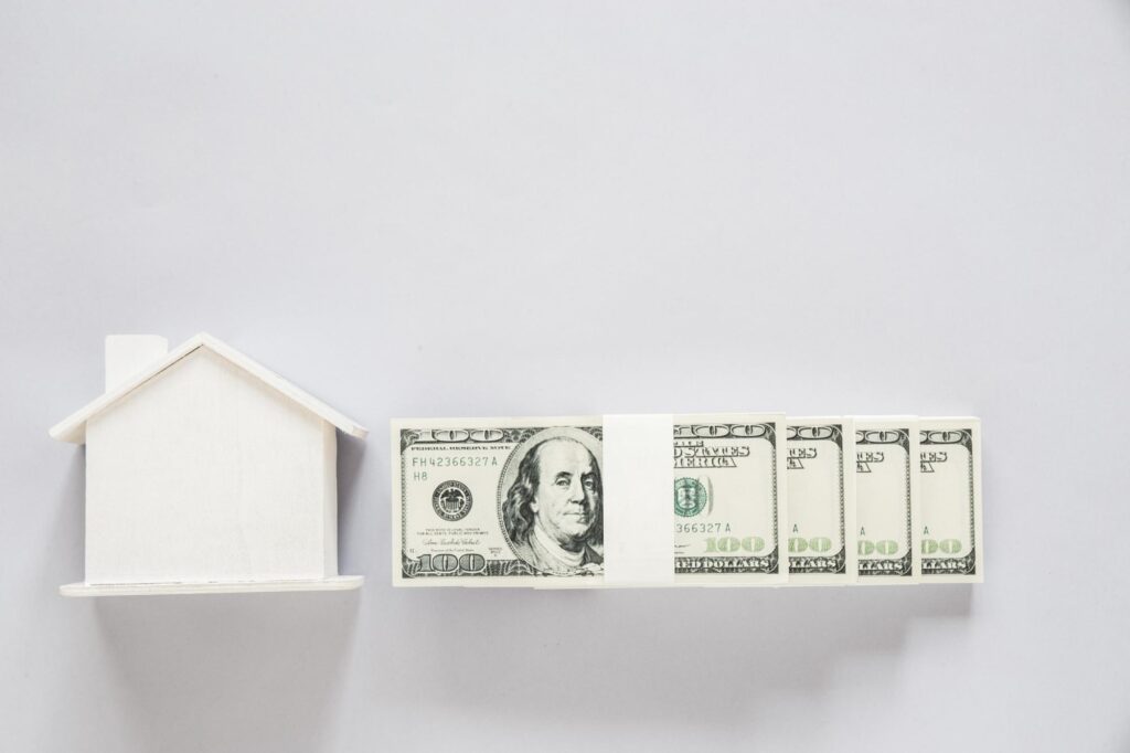 Minimal white model house next to rolled hundred-dollar bills on a white background