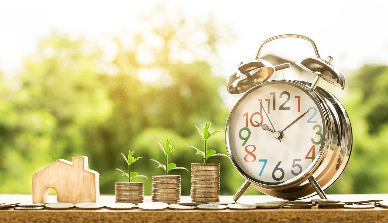 Wooden house model beside stacked coins with seedlings and an alarm clock