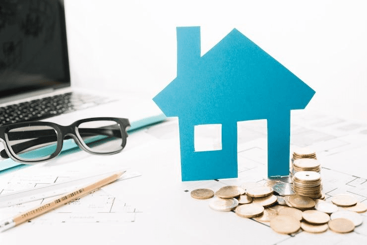 Small house figurine with stacked coins and an open laptop in the background.