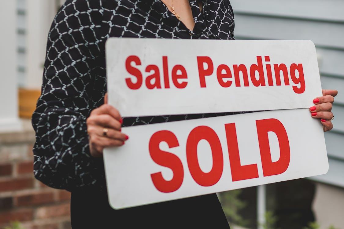 a realtor holding two white and red signages saying “sale pending” and “sold.”