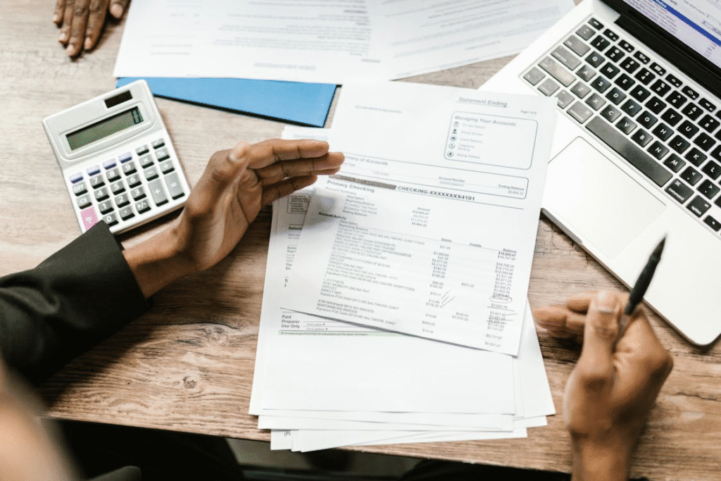 a person looking through bank statement papers placed on a desk with a calculator and a laptop next to them