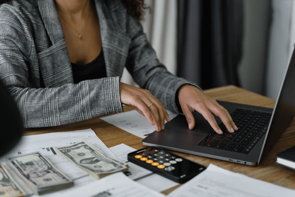 Focused woman in a plaid blazer working on her laptop with documents, calculator, and money lying beside her