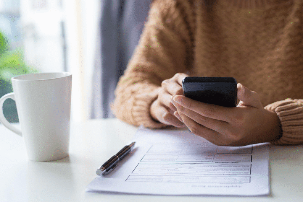 a person going through their phone while filling out a form placed on their desk next to a white cup