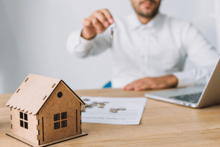 a man holding house keys sitting at a desk with coins and a miniature house model