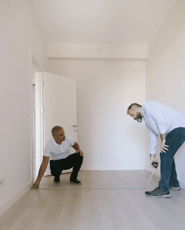 two men in a room with white walls, measuring it up using a measuring tape