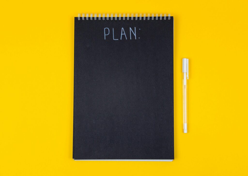 A black notebook on a yellow background with a pen next to it, with “PLAN” written on it
