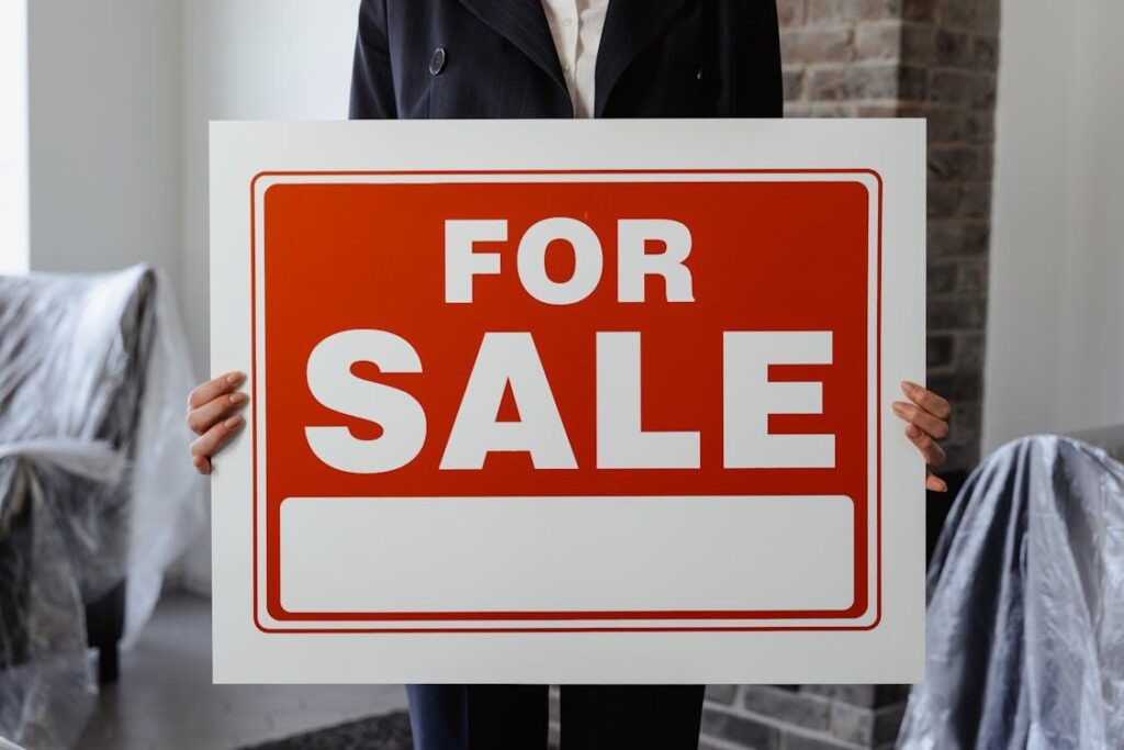 a woman holding a placard that says “for sale” with furniture covered in plastic behind her