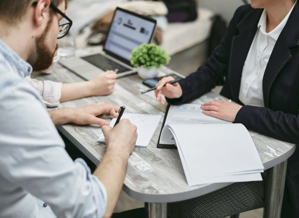 The image shows a collaborative work setting where three individuals are seated around a table, engaging in a meeting or brainstorming session. 