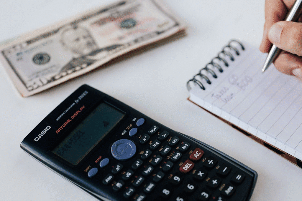 The image shows a calculator, a notepad with handwritten notes, and a dollar bill, suggesting financial calculations or budgeting.