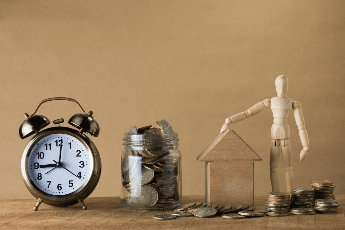 The image features a clock, a jar filled with coins, and a wooden figure placing a coin on a house-shaped structure made of coins.