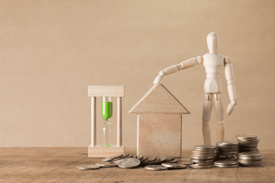 The image depicts a wooden mannequin next to a house-shaped block, coins, and an hourglass, symbolizing themes of time, money, and investment.