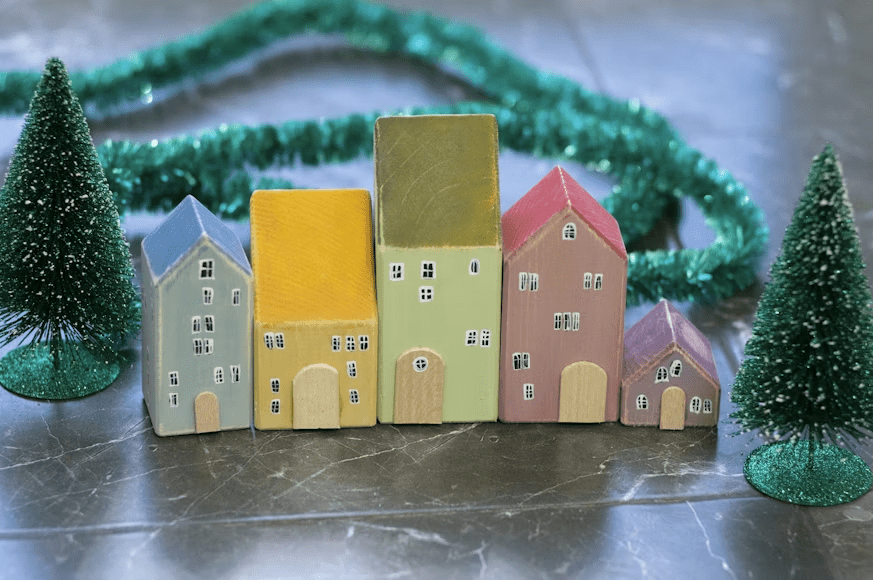 The image shows colorful wooden house figurines arranged with small green Christmas trees and a sparkly garland.