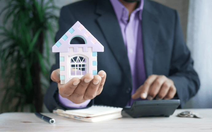 This photo shows a man holding a small house with a notebook, pen, and calculator.