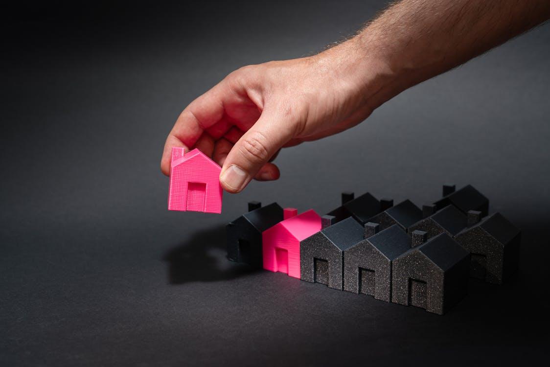 a person holding a pink miniature house among other miniature houses on a black surface