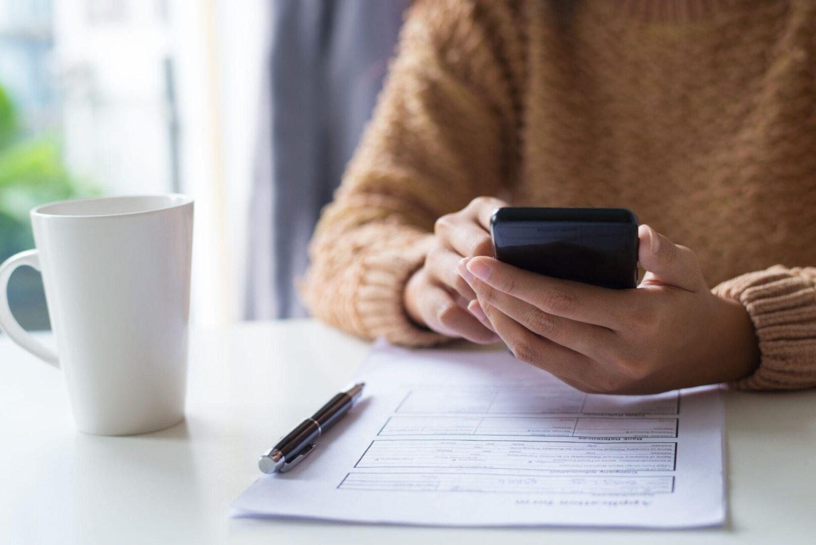 a person going through their phone while filling out a form placed on their desk next to a white cup