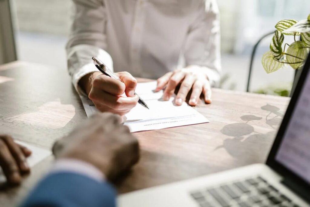 a person sitting across from another person at a desk, signing a form with a pen