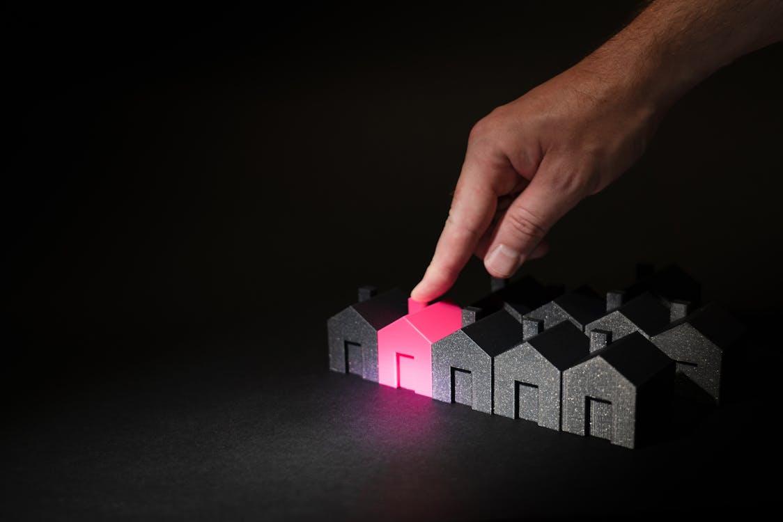 A hand points to a glowing pink miniature house among a row of dark model houses.