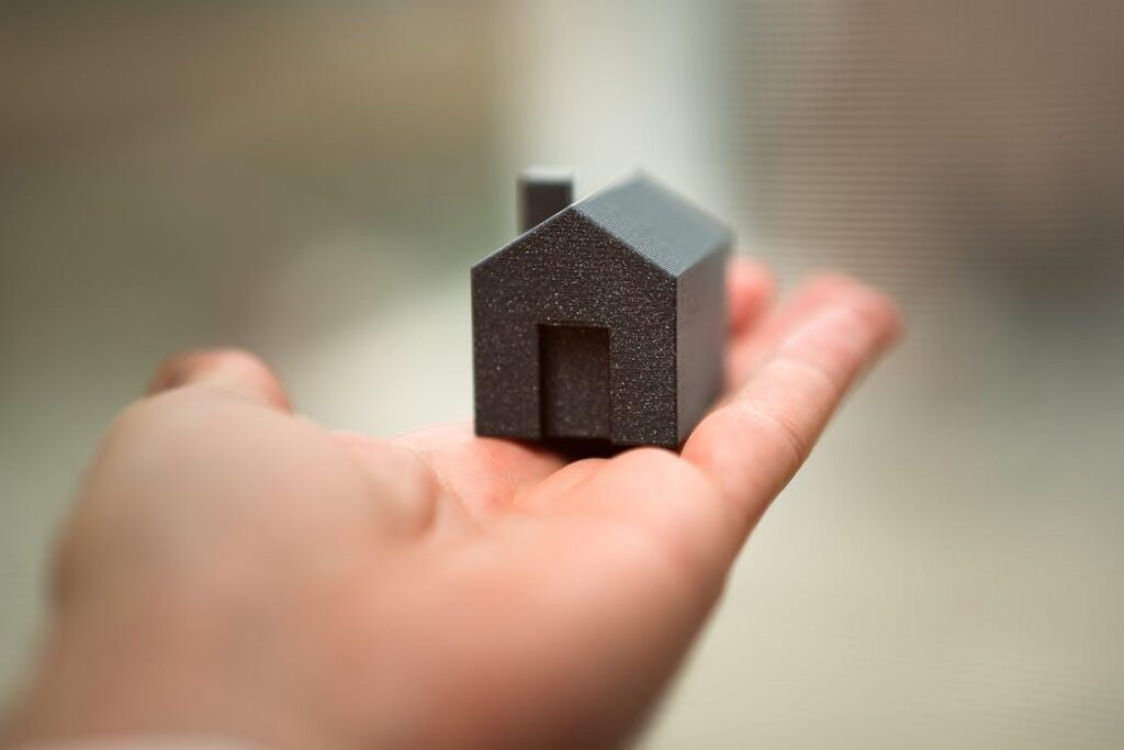 The image shows a hand holding a small, dark-colored house model, symbolizing homeownership or a real estate concept.