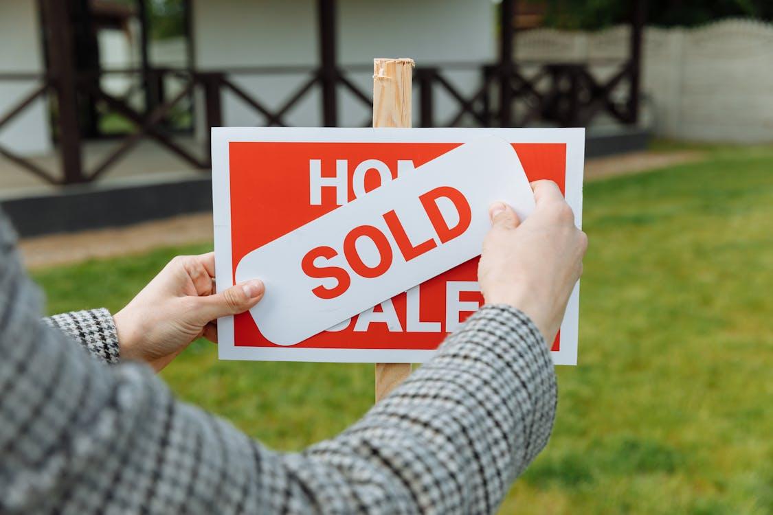 The image shows someone placing a "SOLD" sign on top of a "For Sale" sign in front of a house, indicating the property has been sold