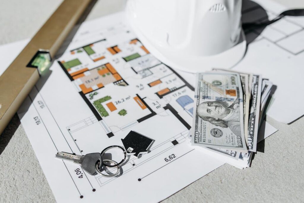 Image of a floor plan laid on a surface with a stack of cash, keys with a house keychain, a ruler, and a construction hat resting on top of it