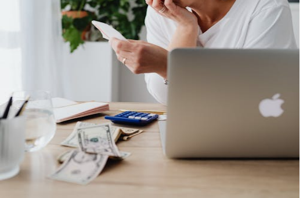 A woman sits at a desk looking worried while checking bills and cash beside a laptop and calculator.