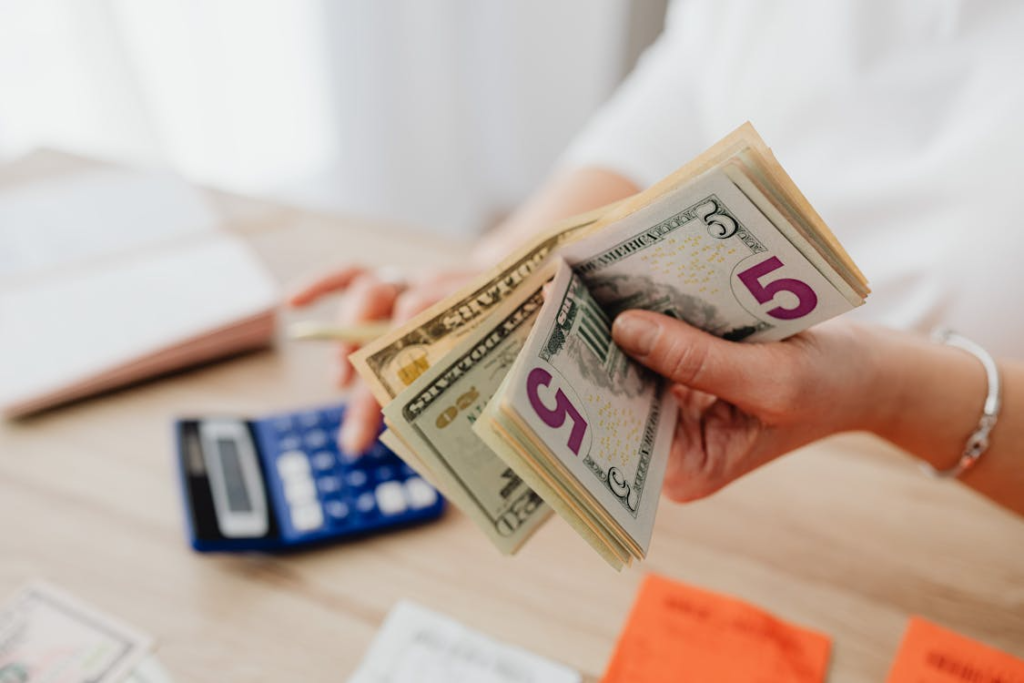 A person holding and counting U.S. dollar bills at a desk with a calculator and paperwork.