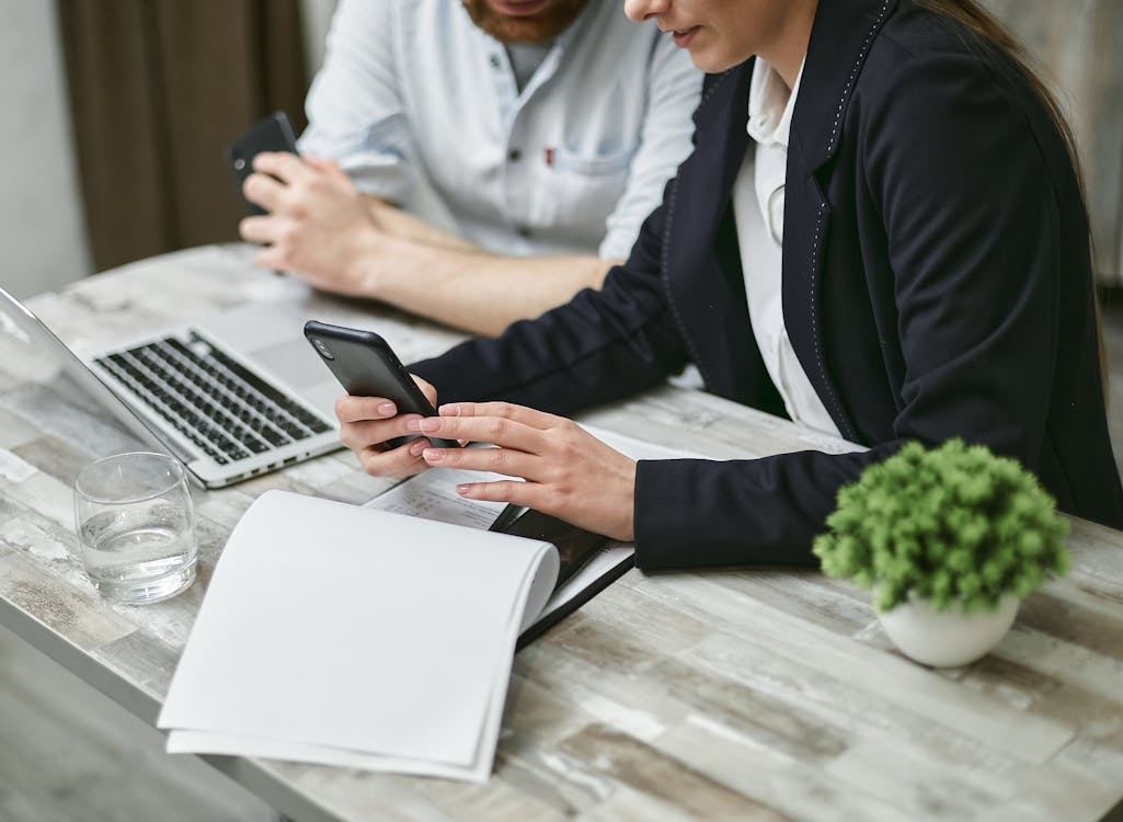 Two professionals working at a desk, using smartphones and a laptop while reviewing documents.