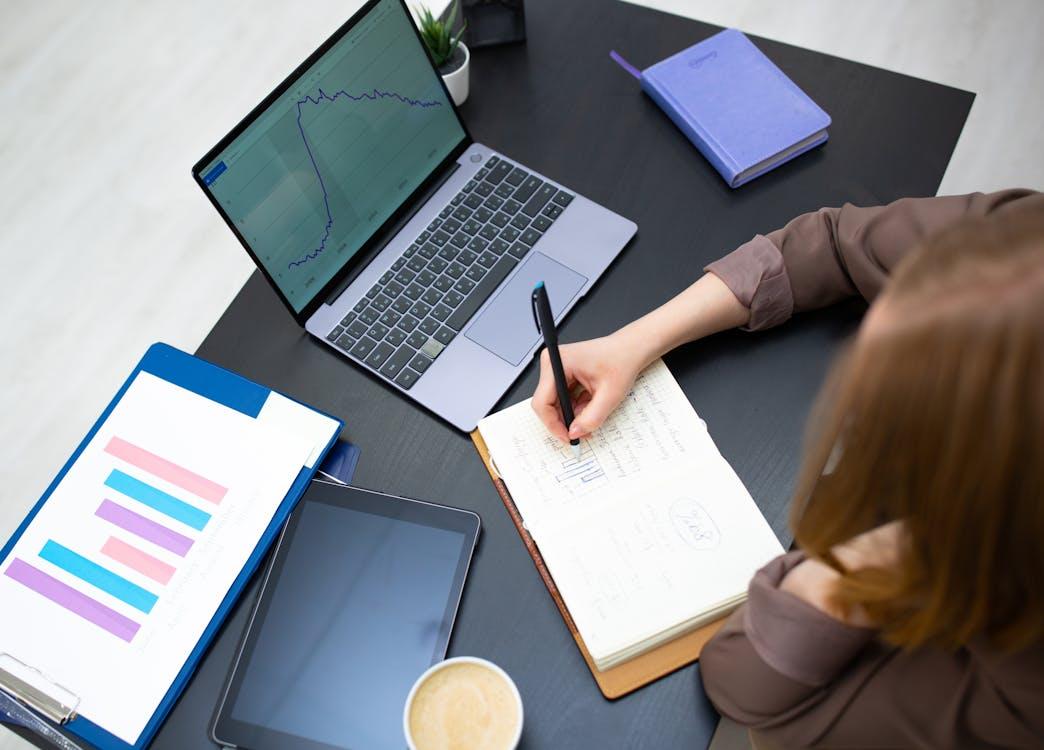 An image of a woman writing in her notebook as she sits in front of a laptop