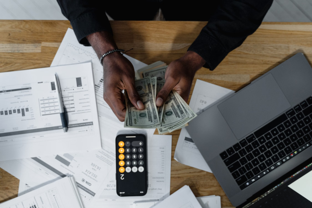 A person counting money with a laptop, mobile with a calculator app, and some papers and a pen on his desk