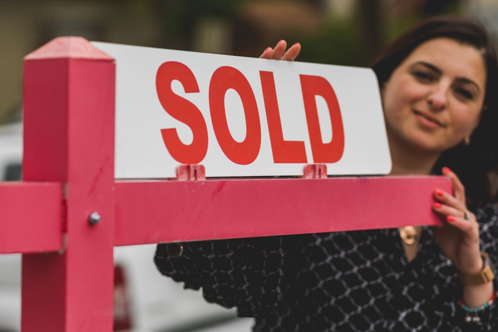 Young woman real estate agent smiling while holding a red For Sale sign outdoors, representing successful property transactions